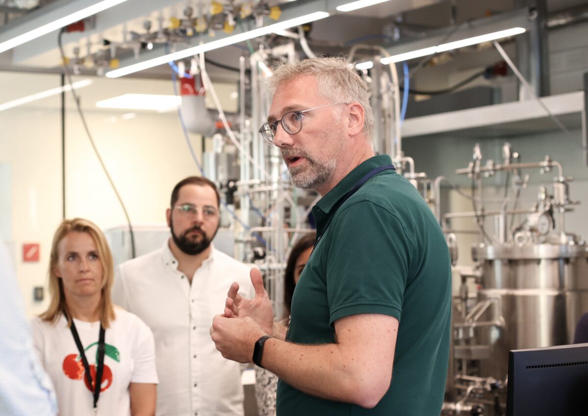 A man in a green polo shirt is speaking to a group of people inside a modern laboratory with stainless steel biomanufacturing equipment. The listeners, some wearing white coats and others in casual clothing, are paying close attention to his explanation.